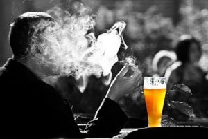 Black and white image of a man in a bar smoking with an order of beer to him. The image is relating to alcohol use among LGBTQQI+.