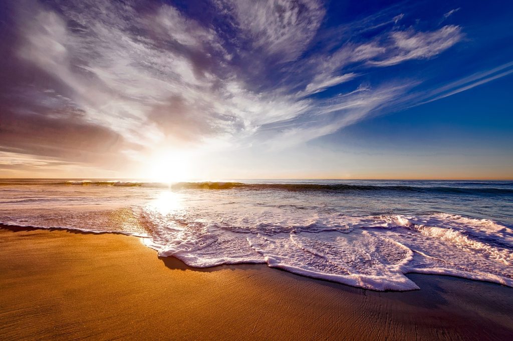 This photo is an image of the beach with blue skies and clouds. A shapeless sun pierces through the clouds and in through the waves and onto a golden sandy shore, which is in the bottom foreground of the picture. The image is utilized due to its ability create a sense of peace, love, happiness, and joy, which are all things that are useful to combat depression.