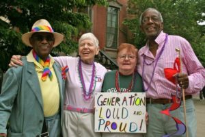 A picture of four aging LGBTQQI* adults. There are two black men and two white women and of the women holds a sign with the words "generation loud and proud!."
