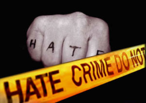 A black and white image of a hand made into a fist with the words "HATE" written on the backside of the fingers with a black marker. There is a yellow and black hate crime tape at the front. It is used for it's clear message that hate is not tolerated against LGBTQQI+.
